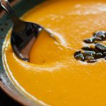 Bildausschnitt einer Kürbissuppe in einem Tonteller mit Löffel und in der Mitte Kürbiskernen